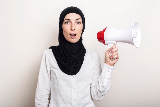 Giovane donna musulmana con una faccia sorpresa in un hijab tiene un megafono nelle sue mani su uno sfondo chiaro banner guarda la telecamera