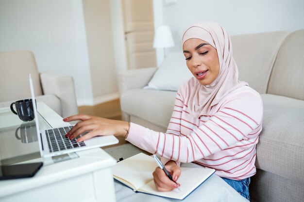 イスラム教徒の若い女性はヒジャブを着て、朝自宅のラップトップで顧客の住所を入力して注文を確認し、コンセプトビジネスを小規模にし、オンラインで買い物をします