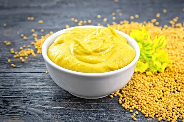 Photo mustard sauce in a white bowl, mustard flower and seeds on a black wooden board background