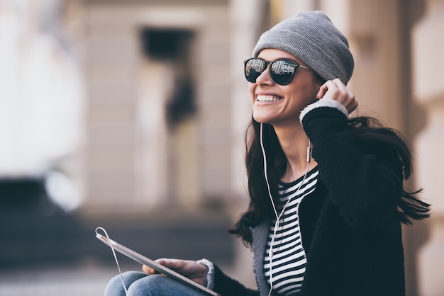 Muziek maakt haar dag. Zijaanzicht van mooie jonge vrouw in zonnebril die haar koptelefoon aanpast en wegkijkt met een glimlach terwijl ze buiten zit