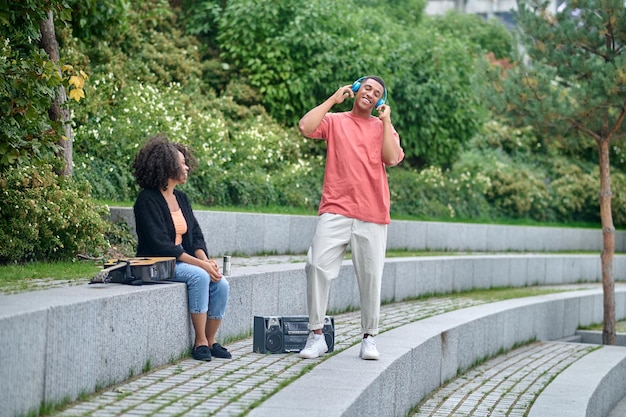 Muziek, plezier. Donkere meid zit te kijken naar staande glimlachende man in koptelefoon met gesloten ogen in groen park