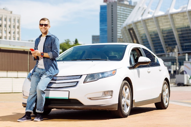 My automobile. Confident smart man standing near his car while holding a smartphone in his hands