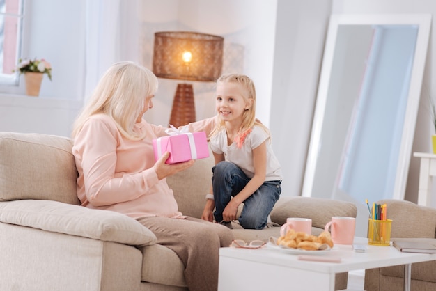 My birthday. Cheerful nice elderly woman sitting on the sofa and feeling happy while receiving a present from her granddaughter