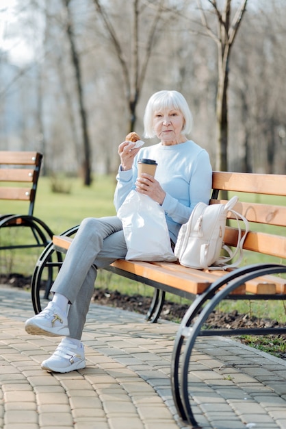 My brunch. Concentrated old woman enjoying her coffee and a cookie while sitting on the bench