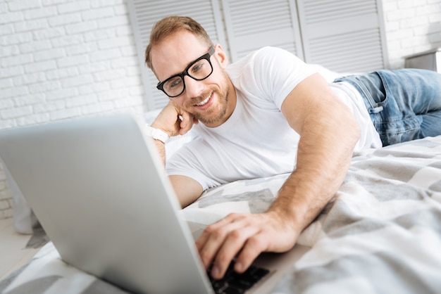 Моя любимая работа. Харизматичный улыбающийся веселый мужчина, лежащий на кровати и использующий ноутбук, выражая интерес и просматривая Интернет