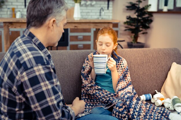 My favourite drink. Nice pleasant girl holding a tea cup while sitting on the sofa with her father