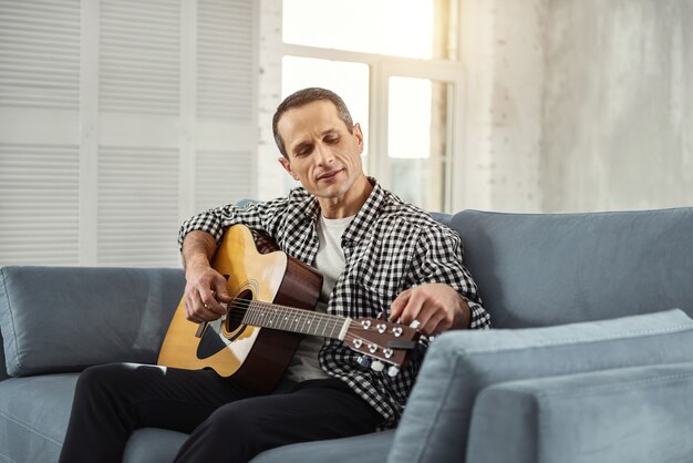 My favourite guitar. Handsome happy well-built man smiling and playing the guitar while sitting on the couch