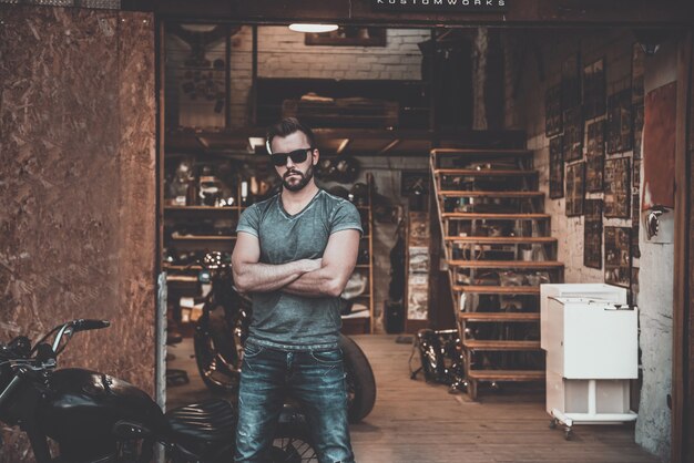 My garage and my rules! Confident young man keeping arms crossed and looking at camera while standing near his motorcycle garage or repair shop