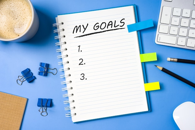 My goals blank list on notebook with the Office equipments and a cup of coffee on blue table background.