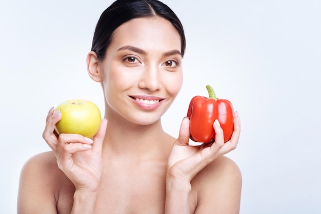 私の健康的な食事。笑顔で赤いピーマンと黄色いリンゴを片手に持っている裸の肩を持つ愛らしい黒髪の若い女性