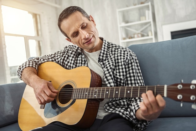 My hobby. Good-looking alert well-built man smiling and playing the guitar while sitting on the couch