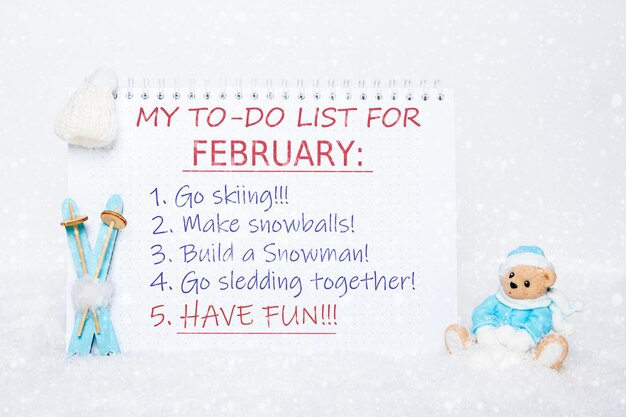 Photo my to-do list for february. notepad with a to-do list: skiing, making snowballs, making a snowman, sledding, having fun and a toy teddy bear in blue clothes, blue skis, a white hat on white snow