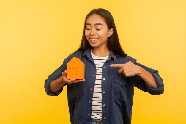 My own home. Portrait of cheerful girl in denim shirt pointing paper house and smiling satisfied, dreaming of real estate purchase, favorable mortgage. indoor studio shot isolated on yellow background