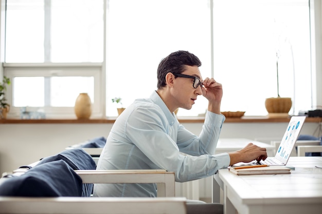 My project. Handsome serious dark-haired man working on his laptop and touching his glasses while sitting at the table