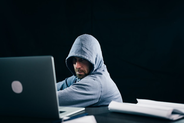 Mysterious bearded man hacker hides his face under the hood, doing something illegal on the laptop