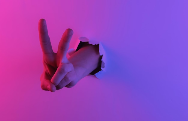 Mystical halloween concept. Hand shows V gesture through a torn hole.
Creative pop art pink blue neon color. Trendy gradient illumination. Night light