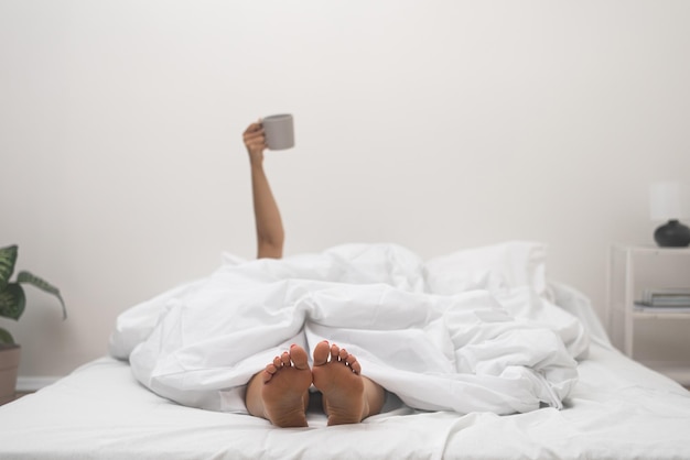 N bed feet escape blanket woman wakes up holding cup of coffee stretching morning