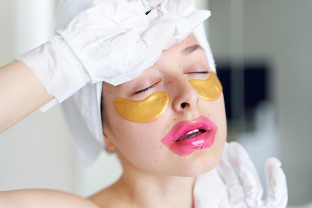 Na een douche gebruikt een meisje gewikkeld in een handdoek cosmetische pleisters voor de huid onder de ogen, lippen en handschoenen om haar handen en voeten te hydrateren. Cosmetische trends voor lichaamsverzorging thuis.