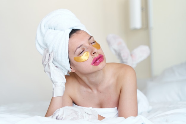 Na een douche gebruikt een meisje gewikkeld in een handdoek cosmetische pleisters voor de huid onder de ogen, lippen en handschoenen om haar handen en voeten te hydrateren. Cosmetische trends voor lichaamsverzorging thuis.