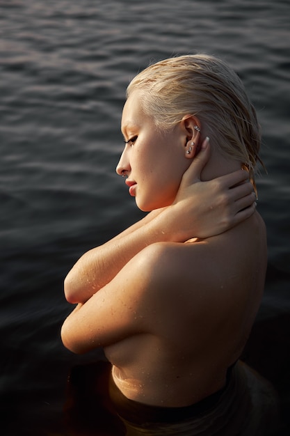 Naakt naakte sexy vrouw in water bij zonsondergang. Mooie blonde vrouw met kort nat haar en grote borsten, kunstportret in zee