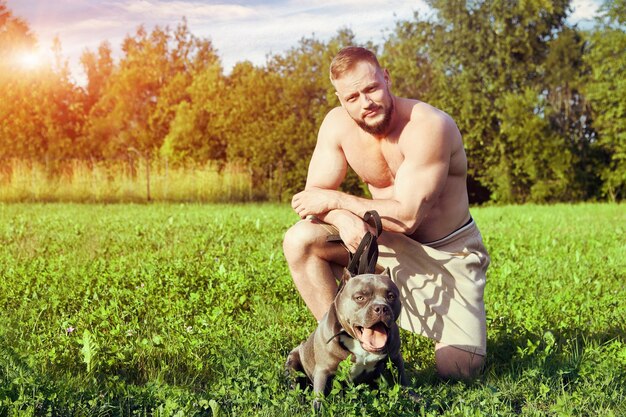 Naakt tot aan de taille baarde man met een Amerikaanse pesthond zit op het gras in het veld leunend op één knie