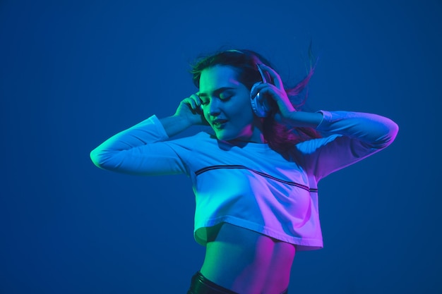 Naar muziek aan het luisteren. portret van de blanke jonge vrouw geïsoleerd op blauwe studio achtergrond in neon licht. mooi vrouwelijk model. concept van menselijke emoties, gezichtsuitdrukking, verkoop, advertentie, jeugdcultuur.