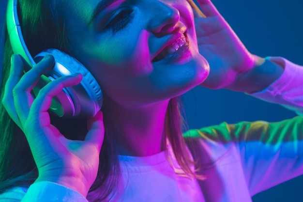 Naar muziek aan het luisteren. Portret van de blanke jonge vrouw geïsoleerd op blauwe studio achtergrond in neon licht. Mooi vrouwelijk model. Concept van menselijke emoties, gezichtsuitdrukking, verkoop, advertentie, jeugdcultuur.