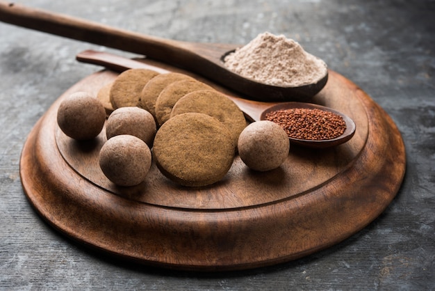 Nachni or Ragi laddu and biscuits or cookies made using  finger millet, sugar and ghee. It's a healthy food from India. Served in a bowl or plate with raw whole and powder