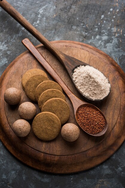 Nachni or Ragi laddu and biscuits or cookies made using  finger millet, sugar and ghee. It's a healthy food from India. Served in a bowl or plate with raw whole and powder