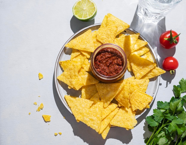 Nachos or tortilla with spicy tomato sauce on a plate with fresh herbs on a white background with shadow Corn chips with salsa sauce