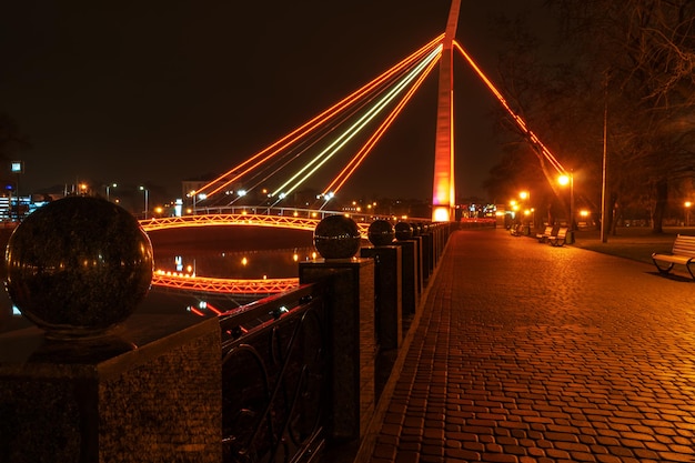 nachtstadslandschap - dijk met straatstenen en een lichtgevende brug met weerspiegeling in de rivier