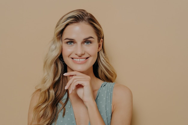 Nadenkend aantrekkelijke jonge blonde vrouw draagt blauwe jurk hand in hand onder de kin, glimlachend en kijkend naar camera met blije gezichtsuitdrukking terwijl ze geïsoleerd naast oranje achtergrond staat