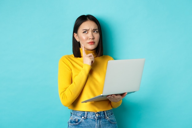 Nadenkend aziatische vrouw die aan laptop werkt, en weg denkt die, besluit neemt, die zich tegen blauwe achtergrond bevindt.