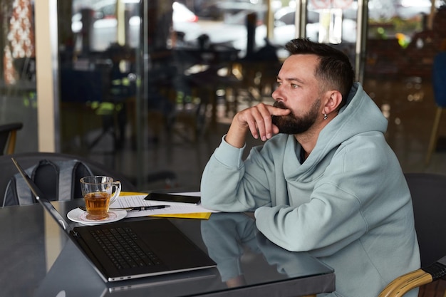 Nadenkend bebaarde man in casual kleding in café met zijn laptop