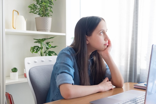 Nadenkend brunette meisje in denim shirt om thuis te zitten studeren schrijven huiswerk communiceert internet