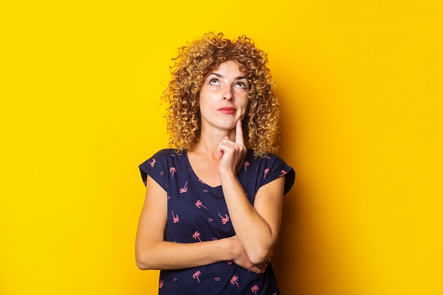 Nadenkend gekrulde jonge vrouw opzoeken met haar vinger tegen haar kin op een gele ondergrond