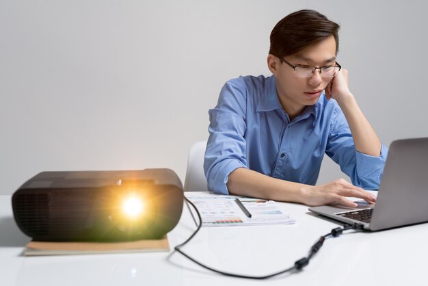 Nadenkend jonge aziatische analist in brillen zit aan bureau met projector en laptop gebruikt tijdens het werken aan presentatie