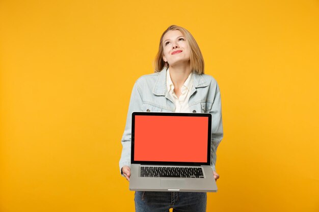 Nadenkend jonge vrouw in denim casual kleding opzoeken, met laptop pc-computer met leeg leeg scherm geïsoleerd op geeloranje achtergrond in studio. Mensen levensstijl concept. Bespotten kopie ruimte.