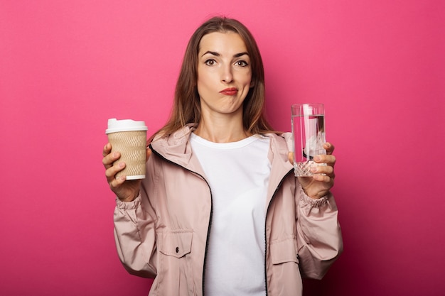 Nadenkend jonge vrouw met papieren beker en glazen beker met water op roze oppervlak