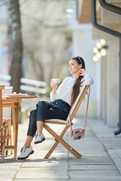 Nadenkend jonge vrouw zittend op het terras van het café in zonnige dag en koffie drinken.