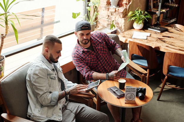 Nadenkend jongeman met behulp van digitale tablet tijdens het plannen van reis met vriend in coffeeshop