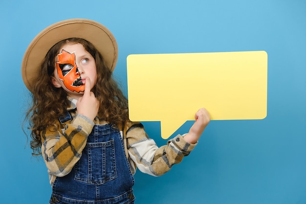 Nadenkend klein meisje met halloween-make-upmasker met een lege tekstballon, denkt na over een beslissing, denkt aan iets, geïsoleerd op een blauwe achtergrond met kopieerruimte. hmm iets vreemds