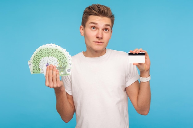 Nadenkend man in witte t-shirt met eurobiljetten en creditcard denken over voordelen van banklening consument nadenken aanbetalingspercentage twijfelen indoor studio shot geïsoleerd op blauwe achtergrond