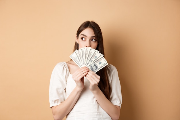 Nadenkend meisje dollarbiljetten houden en lege ruimte kijken, denken wat te kopen op geld, winkelen plannen, staande op beige achtergrond.
