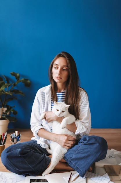 Nadenkend meisje zittend op de vloer met schetsen die witte kat omhelst terwijl ze dromerig opzij kijkt naar een gezellig huis