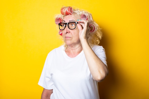 Nadenkend oude vrouw met krulspelden in glazen kijkt naar de zijkant op een geel oppervlak