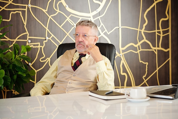 Nadenkend senior ondernemer zittend aan tafel met geopende laptop en kopje koffie