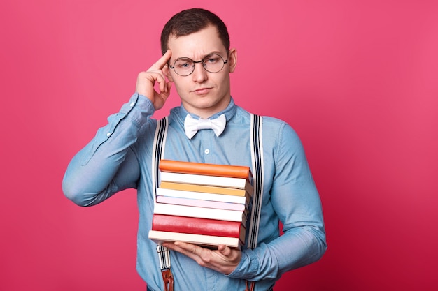 Nadenkend verward druk jong model houdt een stapel boeken in één hand, raakt de tempel aan met de wijsvinger, kijkt opzij, draagt casual kleding, geïsoleerd over roze