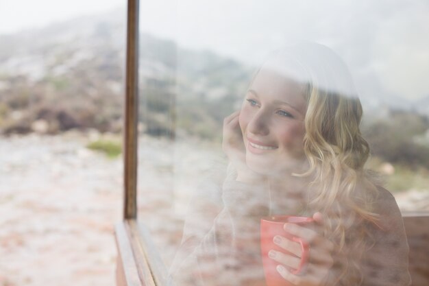 Nadenkende inhoudsvrouw die met koffiekop door venster kijkt