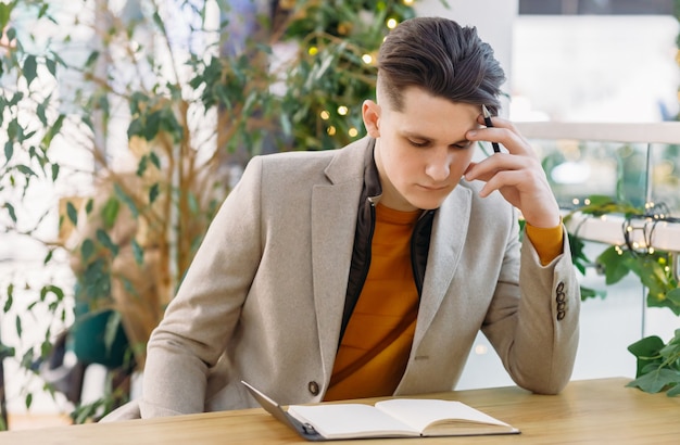 Nadenkende man in beige jas zittend aan een tafel met een pen in zijn handen in een café